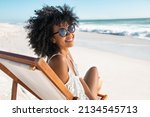 Portrait of happy young black woman relaxing on wooden deck chair at tropical beach while looking at camera wearing spectacles. Smiling african american girl with fashion sunglasses enjoying vacation.