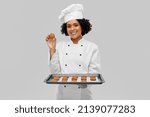 cooking, culinary and bakery concept - happy smiling female chef or baker in white toque and jacket holding baking tray with oatmeal cookies over grey background