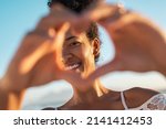 Small photo of Close up of hands of young woman making heart shape. Portrait of a lovely black woman making heart with fingers while looking at camera. Smiling african american woman making love sign during sunset.