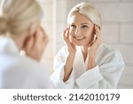 Small photo of Gorgeous mid age adult 50 years old blonde woman standing in bathroom after shower touching face, looking at reflection in mirror smiling doing morning beauty routine. Older dry skin care concept.