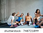 Friends in sportswear high fiving and laughing together while sitting on the floor of a gym after a workout