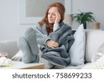 Sick woman with a headache sitting on a sofa at home wrapped in grey blanket
