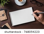Top view of businessman writing on blank notebook. High angle view of man hand writing on empty notepad with spectacles, coffee cup and folder on wooden table. Closeup of male  hand making notes.
