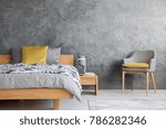 Grey chair and lamp on wooden nightstand in dark bedroom with concrete wall and yellow pillow on bed
