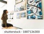 Small photo of Side view of a beautiful young woman studying and watching the abstract paintings and colorful canvases during a visit to the art gallery