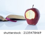 Small photo of Red bitten apple with an open Holy Bible Book on white background. The Christian biblical concept of the biblical story in Genesis of forbidden fruit, temptation, and disobedience to God. A closeup.