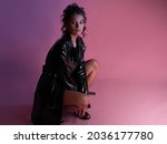 Stylish young brunette in a latex raincoat over a black bodysuit, fashion image on a pink background. Beautiful young female model squatting