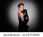 Small photo of A femme fatale in a black dress with an open back and long velvet gloves. Attractive young blonde with makeup for a celebration, portrait in a spot of light