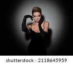 Small photo of A femme fatale in a black dress with an open back and long velvet gloves. Attractive young blonde with makeup for a celebration, portrait in a spot of light