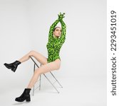 Small photo of Stylish bright dancer in a bright green leopard print bodysuit, posing on a chair, studio photo on a white background. Plastic body, pretentious pose