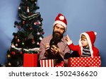 Small photo of Man and boy in Santa hat and hood play with puppies. Pet for Christmas concept. Father and son with strict faces unpack presents on blue background. Dad with beard and kid hold dog near Christmas tree