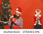 Small photo of Man and boy in Santa hat play hide and seek with puppy. Father and son unpack present boxes on red background. Dad with beard and kid hold little dog near Christmas tree. New year gift concept.