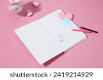 Pink background featured an opening notebook with origami hearts and colorful crayons. A wine glass filled with marshmallows displayed. Copy space for Valentine or Women's day content