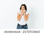 Small photo of Portrait of frightened young woman biting her fingers in horror, looking around warily, white studio background