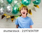 Small photo of boy celebrates his birthday. the child stands against the wall with balloons and squeals. a schoolboy screams next to a wall decorated for the holiday.