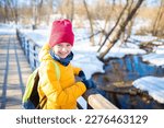 smiling school boy with a backpack in bright clothes goes to school, stands on a bridge in a winter snowy sunny park