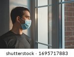 Quarantine self-isolation. Sad young man in a medical mask who looks out the window through the window. Infected man in medical mask on self-isolation looks at the street through the window of a house