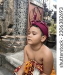 Small photo of 4 years old boy with cranky gesture Wearing balinese traditional clothes and hat sitting on the gate of the church in Bali Indonesia