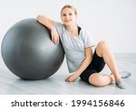 Fitness equipment. Sport indoors. Active kids. Training essentials. Gymnastic accessories. Smiling happy athletic girl sitting on floor with gym ball on light background.
