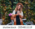 Curly woman posing with vintage bicycle and cup of coffee near green plant wall. Female wearing brown hat and cardigan is travelling by bike with big basket of pink camomiles. Concept of leisure.