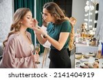 Small photo of Beauty specialist applying eyeshadow on client eyelid in visage studio. Young woman sitting at dressing table and holding glass of champagne while makeup artist doing professional makeup.