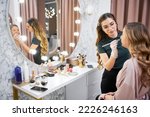 Small photo of Young woman sitting at dressing table and looking in the mirror while makeup artist applying concealer with cosmetic brush. Female stylist doing professional makeup for client in beauty salon.