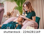 Small photo of Female eyelashes technician in protective face mask combing woman lashes with disposable mascara brush in beauty salon. Beauty specialist comping client eyelashes after extension procedure.