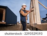 Man worker building wooden frame house on pile foundation. Carpenter hammering nail into wooden joist, using hammer. Carpentry concept.
