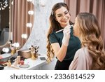 Small photo of Smiling female stylist in glasses applying foundation on client face with cosmetic brush. Joyful beauty specialist doing professional makeup for woman in visage studio
