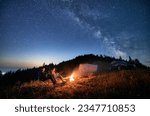 Small photo of Night camping in the mountains under starry sky with Milky way. Travelling couple having a rest at campfire, tourist tent and off-road vehicle in the evening on mountain lawn and stargazing together.