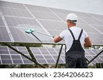 Small photo of Worker cleaning solar photovoltaic panel. Man technician making sure solar batteries in good condition. Back view of professional worker in overalls and helmet cleaning panel by mop.