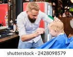 Small photo of Man barber cutting little boy's hair using comb and scissors. Child getting haircut from adult male, likely barber. Professional hairdresser and cute client at modern barbershop.