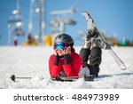 Smiling female skier wearing ski goggles lying with skis on snowy at mountain top and looking away in sunny day with ski lifts and blue sky in background. Close-up