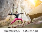 Small photo of Sporty slim woman in pink footless is practicing yoga and doing asana Virabhadrasana 2 outdoors. Huge rocks on the background