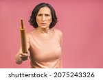 Small photo of A very angry senior mature aged lady holding a rolling pin and threatening to whack someone with it (her husband?). Isolated on pink background.