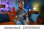 Small photo of Upset bearded hippie man at home couch showing red arrow pointing down, concept of downgrade, unsuccessful business, fall of stock market money exchange rate bankruptcy fail. Hipster guy in night room