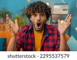 Small photo of Oh my God, Wow. Young indian man surprised looking at camera with big eyes, shocked by sudden victory, good win news, celebrating. Portrait of excited amazed hindu guy at home apartment room on sofa