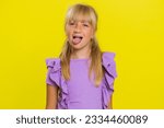 Small photo of Cheerful funny young blonde school girl showing tongue making faces at camera, fooling around, joking, aping with silly face, teasing, bullying, abuse. Preteen female child kid on yellow background