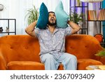 Small photo of Repair work at neighbors. Irritated Indian young man cover ears with pillows annoyed by noisy loud music neighbors suffer from headache wishes silence. Thin walls at home flat without sound insulation