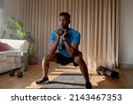 Small photo of Young African American male exercising from home concentrating while squatting with a dumbbell in living room, workout at home