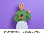 Small photo of Excited young Asian woman in green sweater and hijab holding mobile phone and pointing finger up, creating genius solutions isolated over purple background