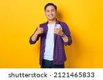 Small photo of Smiling young Asian man in plaid shirt holding mobile phone and making thumb up gesture, approve good thing isolated on yellow background