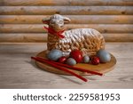 Easter lamb cupcake with red ribbon, painted gray and red eggs and church candles on a vintage board in a wooden house on the table. Traditional festive food for Easter.