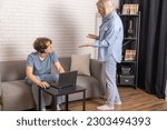 Small photo of Internet addiction. Woman scolding her son while he using laptop in living room the whole day. Mother hysterically trying to reach out to indifferent son, awkward age problems