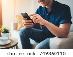 Happy smiling young man using modern smartphone device while sitting on sofa at home, modern design interior, cheerful hipster guy typing an sms message at social network
