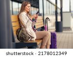 A young and beautiful woman with a suitcase and a backpack is sitting at the train station waiting for a train while using a mobile phone.
