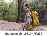 A young woman with curly hair in her 20s exploring the forest trail during autumn, wearing warm clothing and equipped with a yellow backpack, trekking poles, and binoculars.