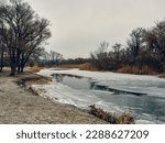 Small photo of The forest river is ice-bound, however, due to the thaw in places, the ice thawed and a polynya (thawed patch) formed, grass and reed trees grow on the banks of the river, cloudy