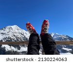 Woman's legs in red ski boots resting on wooden fence after a ski tour on sunny day, snowy mountains in the background. High quality photo