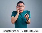 Small photo of Shocked young Asian man in casual t-shirt using mobile phone, reacting to new app isolated on grey background. indonesian independence day celebration concept